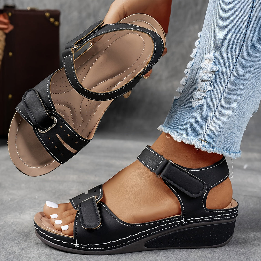 Sandales compensées noires à brides auto-agrippantes et coutures blanches, portées avec un jean effrangé, l'autre sandale tenue en main pour voir la semelle. 4