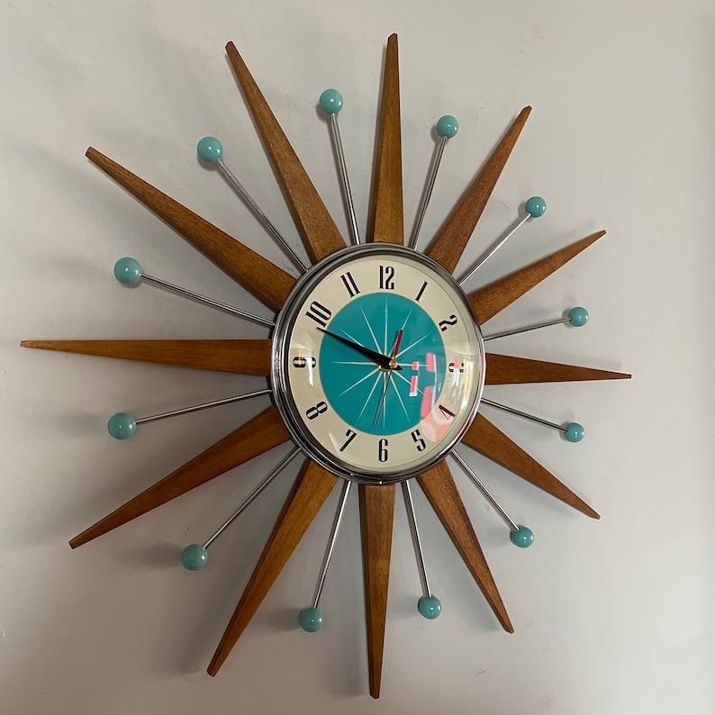 Horloge soleil accrochée à un mur gris clair, vue de face, rayons en bois et boules turquoise au bout des tiges. 2