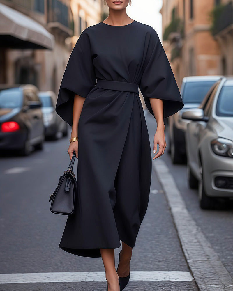Robe noire mi-longue ceinturée à larges manches, portée en pleine hauteur dans une rue avec un sac à main noir et des escarpins. 1