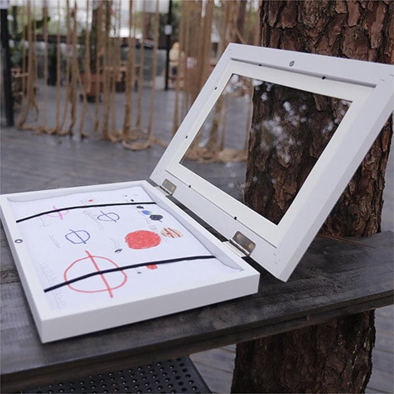 Cadre blanc posé sur une table en bois, porte vitrée relevée, dessin d'enfant maintenu par deux bandes élastiques. 20