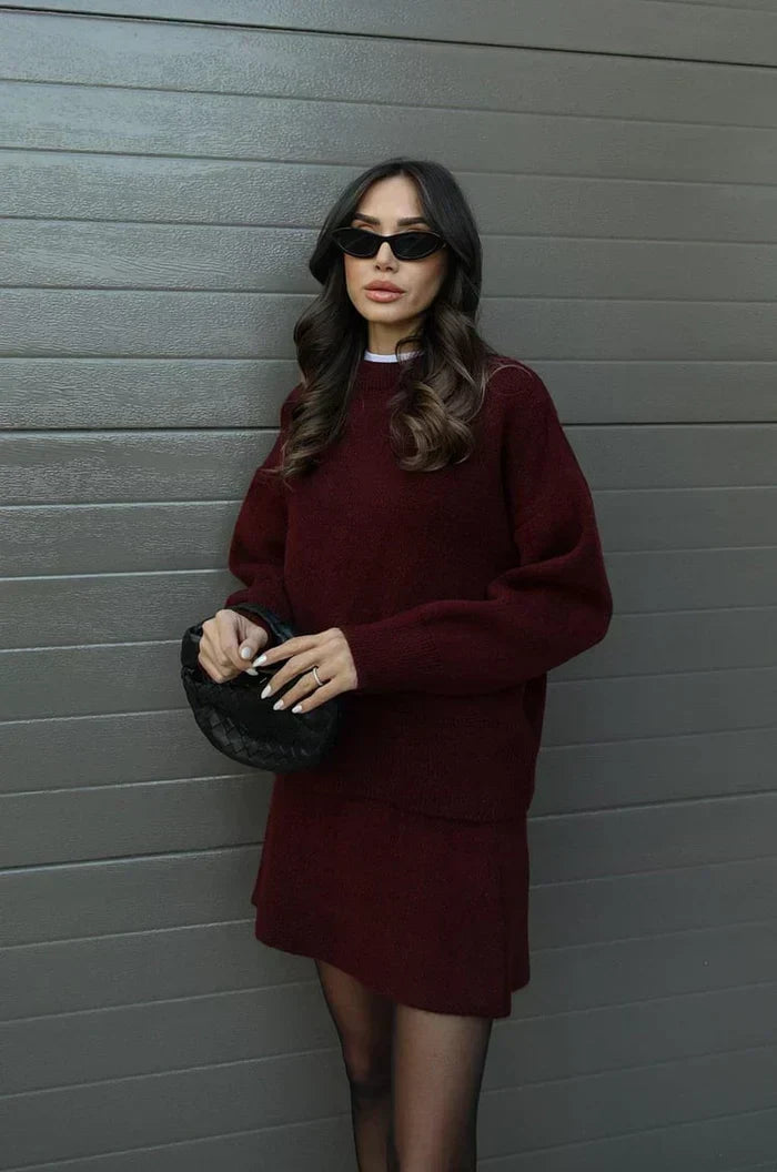 Mannequin en tenue bordeaux, pull à manches longues et jupe courte, appuyée de côté contre un mur gris, sac noir en main. 3