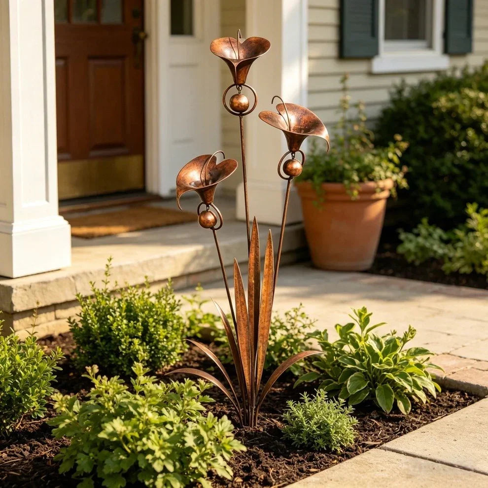 Sculpture de jardin en cuivre vieilli avec trois fleurs en cloche, plantée dans un massif devant l'entrée d'une maison, près d'un pot en terre cuite. 1