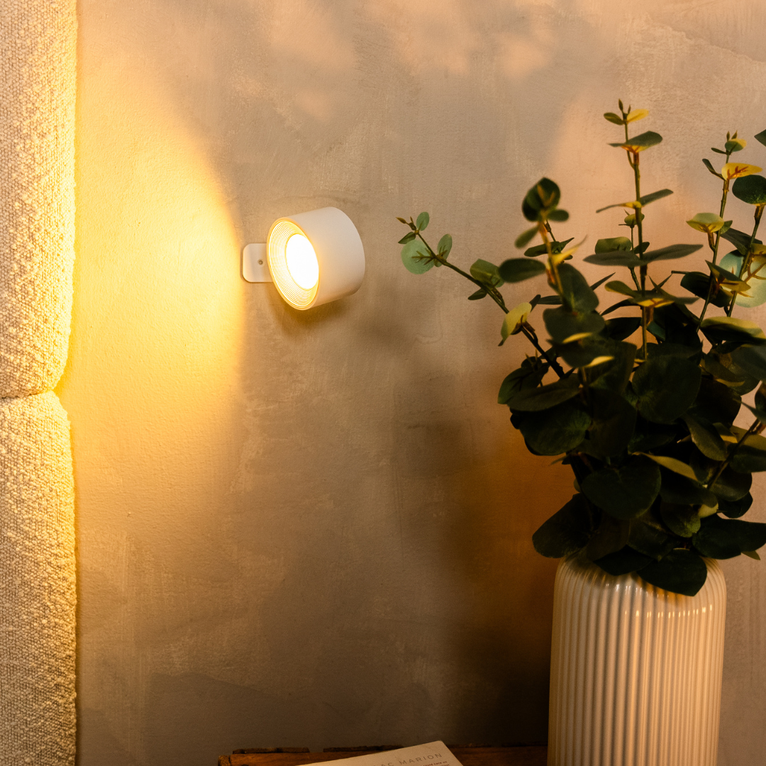 Applique blanche allumée dirigeant sa lumière dorée sur le côté, contre un mur patiné, avec un vase strié et des feuilles d'eucalyptus. 14