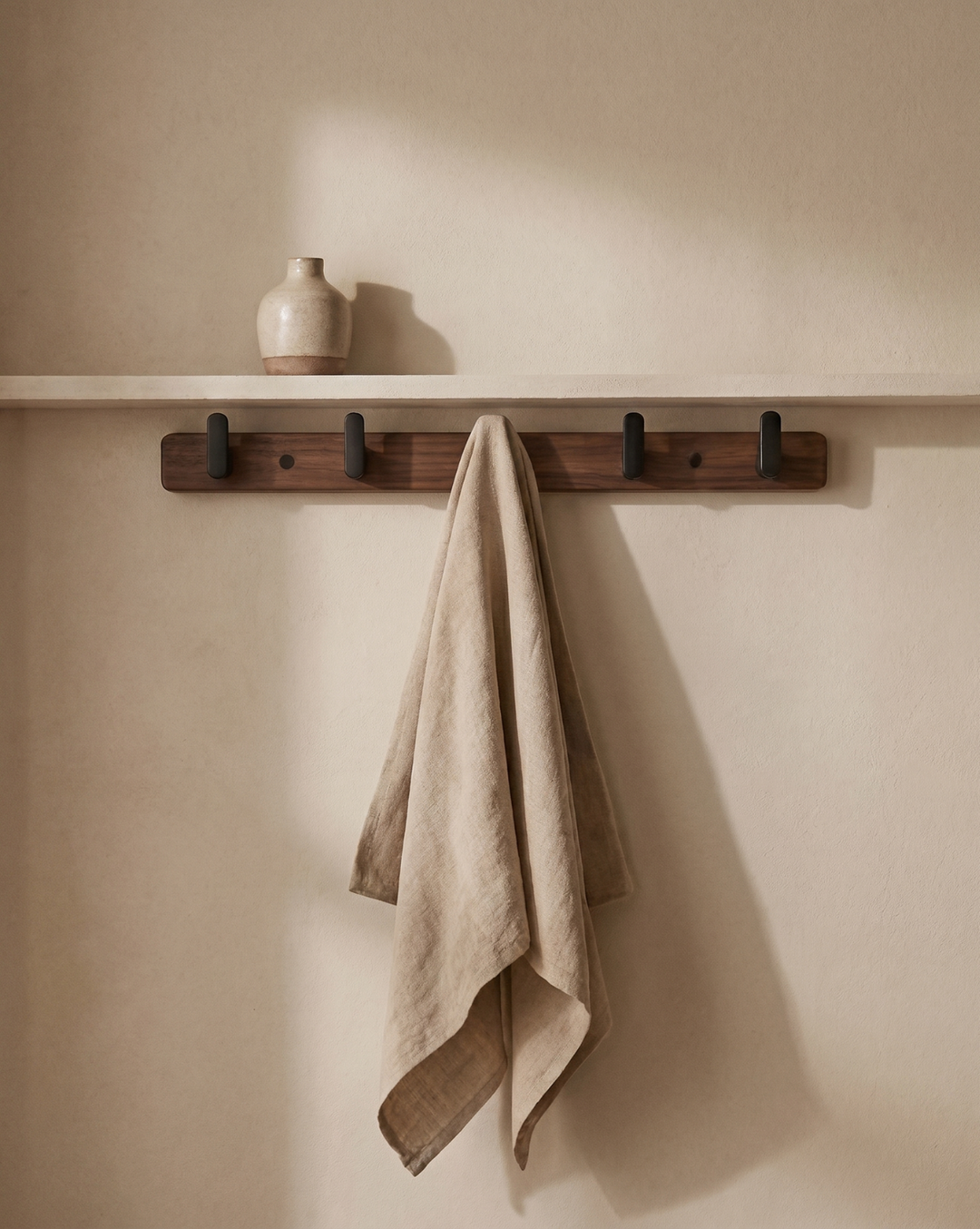 Barre à quatre crochets noirs fixée au mur sous une étagère, avec un linge beige suspendu 1