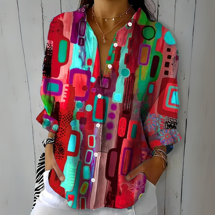 Blouse à motif abstrait rouge, turquoise et violet, portée de face avec colliers fins et pantalon blanc. 4