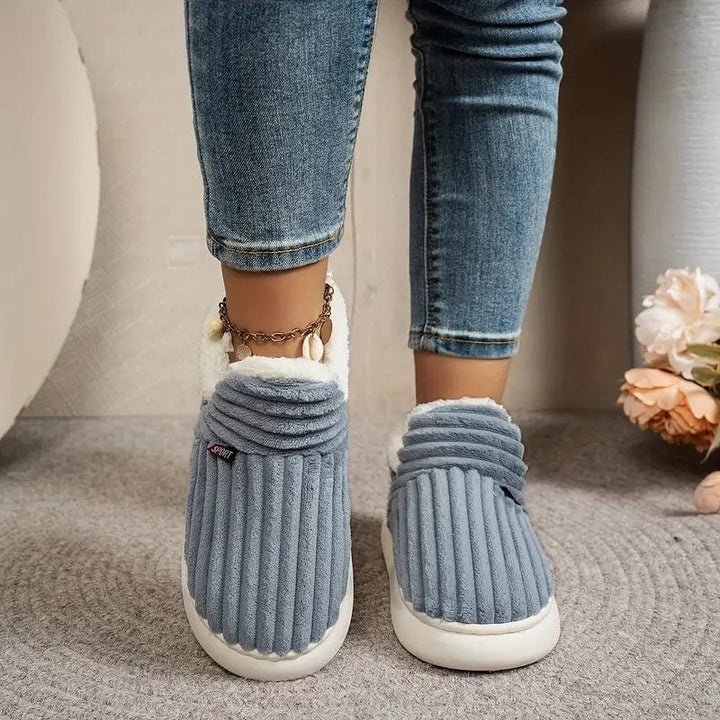 Vue de face de chaussons gris-bleu côtelés portés avec un jean, bord intérieur en peluche blanche visible. 11