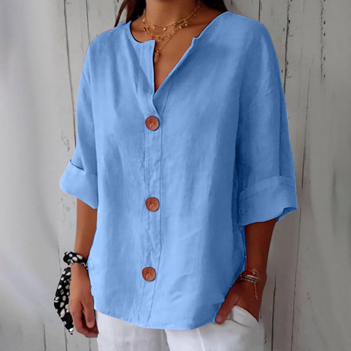 Blouse ample bleu ciel portée par une mannequin, col en V, trois gros boutons en bois et manches retroussées, devant un mur en lattes. 1