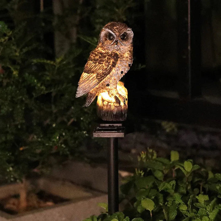 Hibou brun éclairé de nuit sur sa souche lumineuse, planté au milieu de feuillages dans un massif de jardin. 4