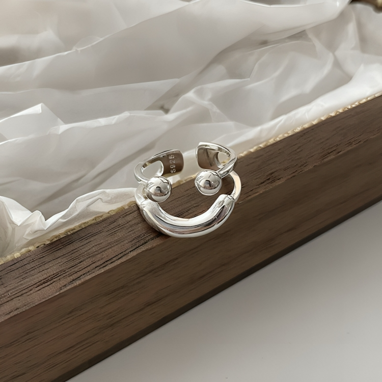 Bague sourire argentée posée sur un plateau en bois sombre, avec du papier de soie blanc froissé en arrière-plan. 6