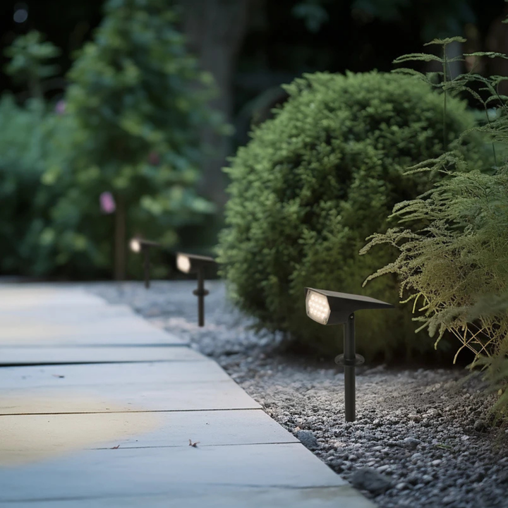 Trois projecteurs solaires plantés en ligne le long d'une allée dallée, bordée de gravier et de buissons, éclairant doucement le jardin à la tombée de la nuit. 1