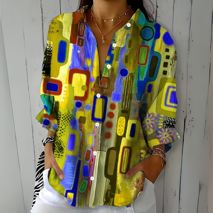Blouse à motif abstrait jaune, bleu et vert, portée de face avec un pantalon blanc et plusieurs bracelets. 5