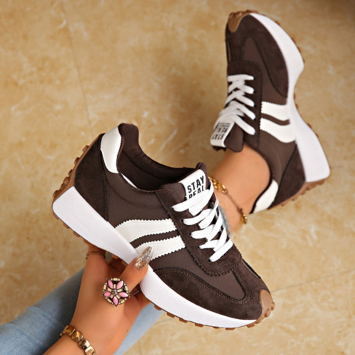 Baskets marron foncé à bandes blanches, tenues en main au-dessus d'un sol en marbre beige 3