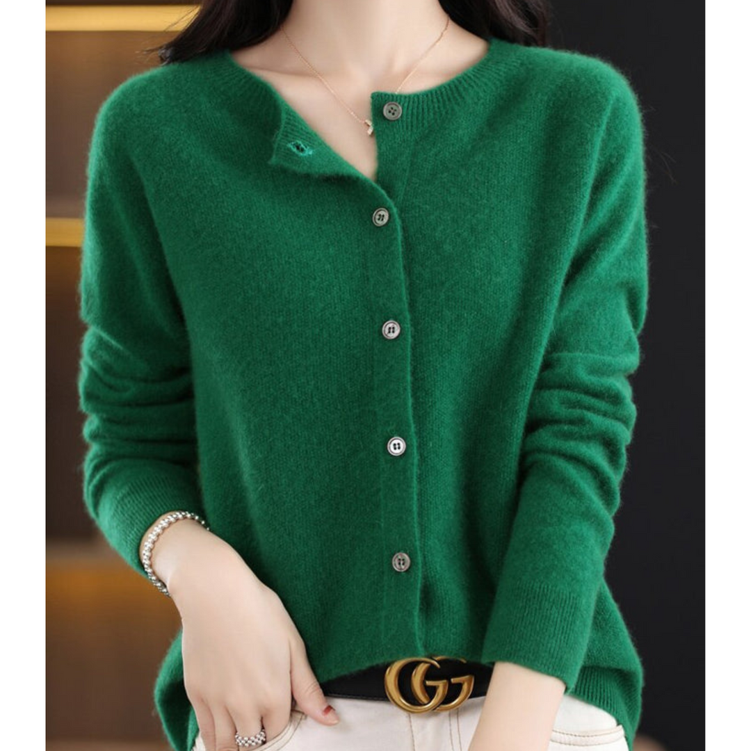 Cardigan vert foncé à col rond et boutons gris, porté avec un pantalon blanc, un bracelet et une ceinture noire. 12
