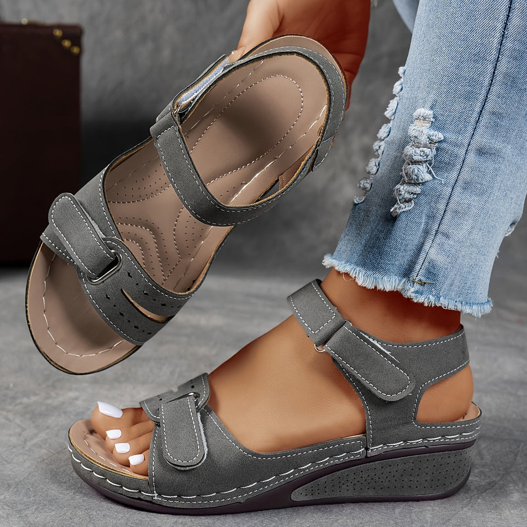 Sandales compensées grises à aspect daim, brides auto-agrippantes et coutures blanches, portées avec un jean, l'autre sandale tenue en main côté semelle. 6