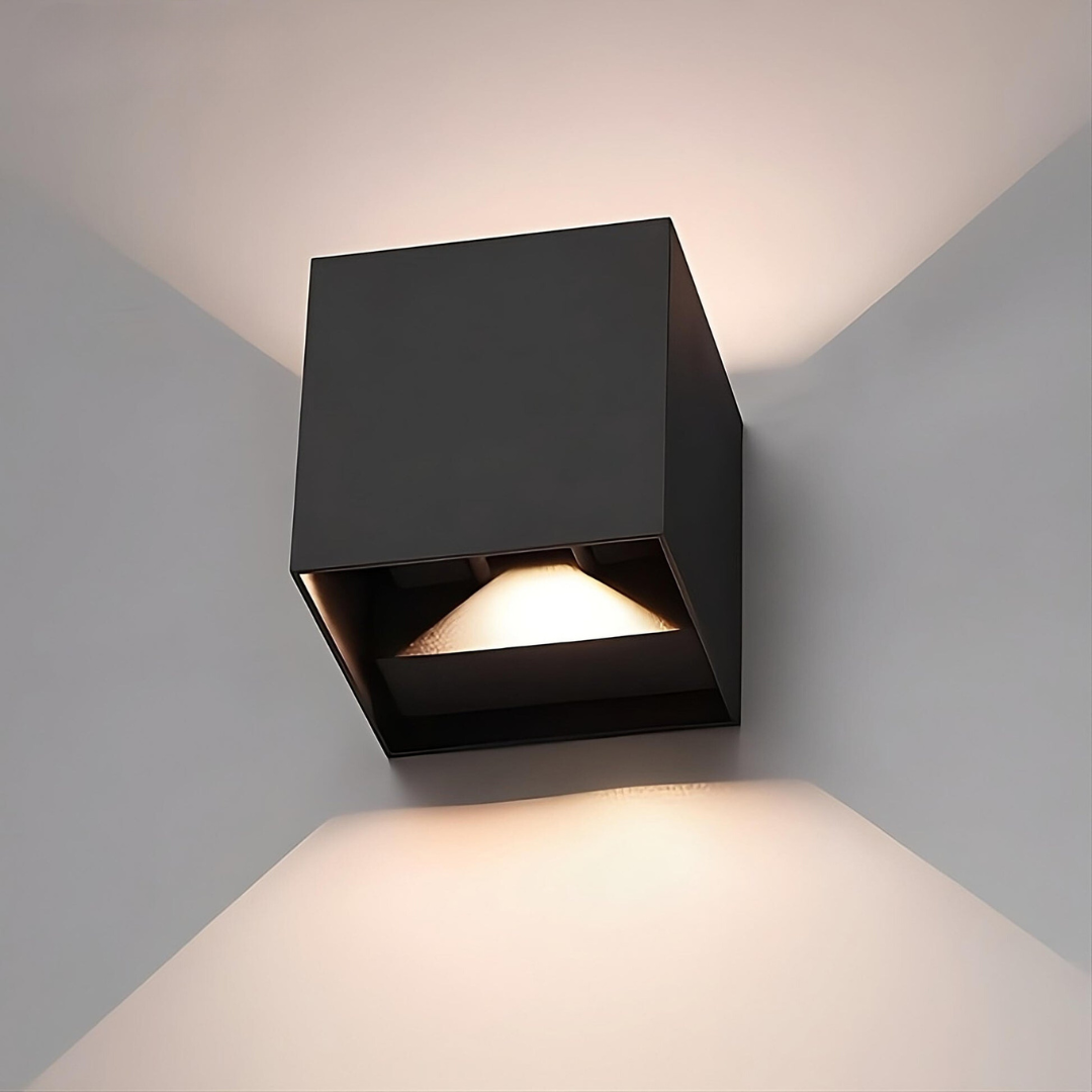 Gros plan sur une applique cubique noire montrant l'ouverture inférieure où la source lumineuse diffuse une lueur chaude sur le mur. 16