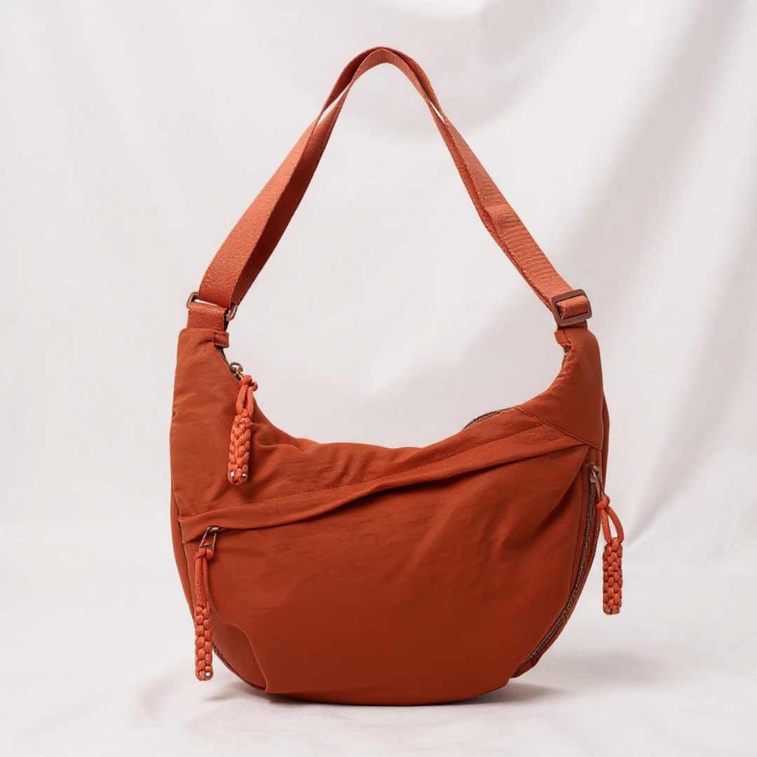 Sac en forme de croissant orange rouille sur fond de tissu clair, avec anse réglable et zips à pompons tressés. 1