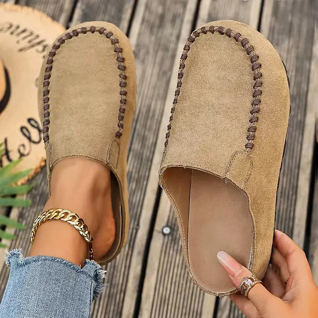 Mules beige façon daim à surpiqûres foncées vues de dessus, l'une portée sur un parquet en bois, l'autre tenue à la main. 2