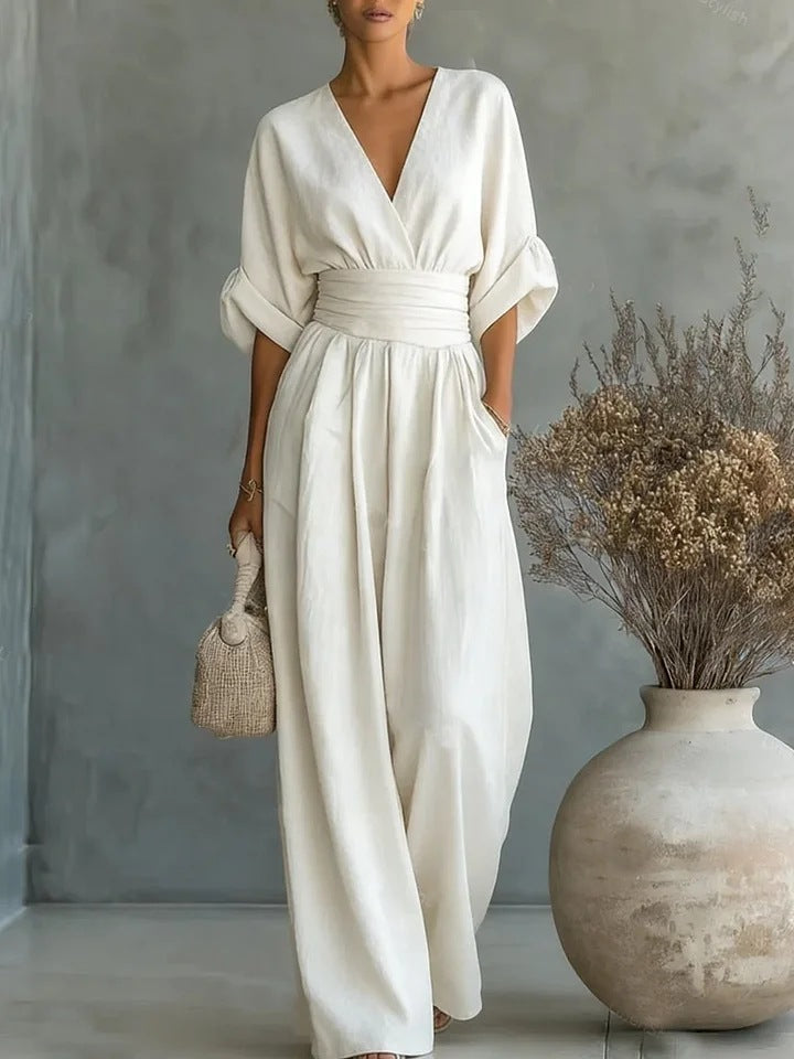 Combinaison blanc crème portée en plan large devant un mur gris, avec un grand vase et des fleurs séchées. 1