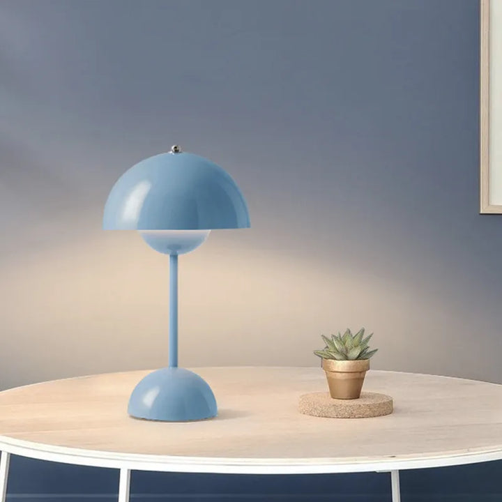Lampe champignon bleu clair sur une table ronde claire, avec un petit cactus en pot doré, devant un mur bleu-gris. 6