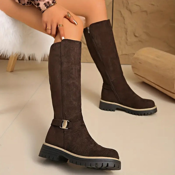 Bottes marron foncé portées assise, vue de face montrant la boucle dorée sur la sangle et la fermeture éclair. 6