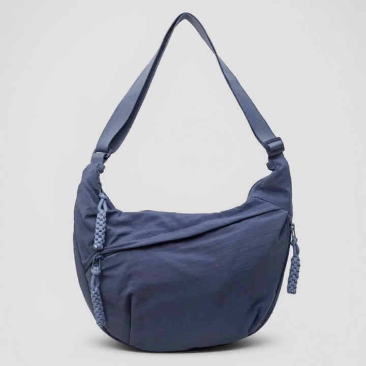 Sac demi-lune bleu en nylon vu de face sur fond clair, avec bandoulière réglable et zips ornés de tirettes tressées. 1