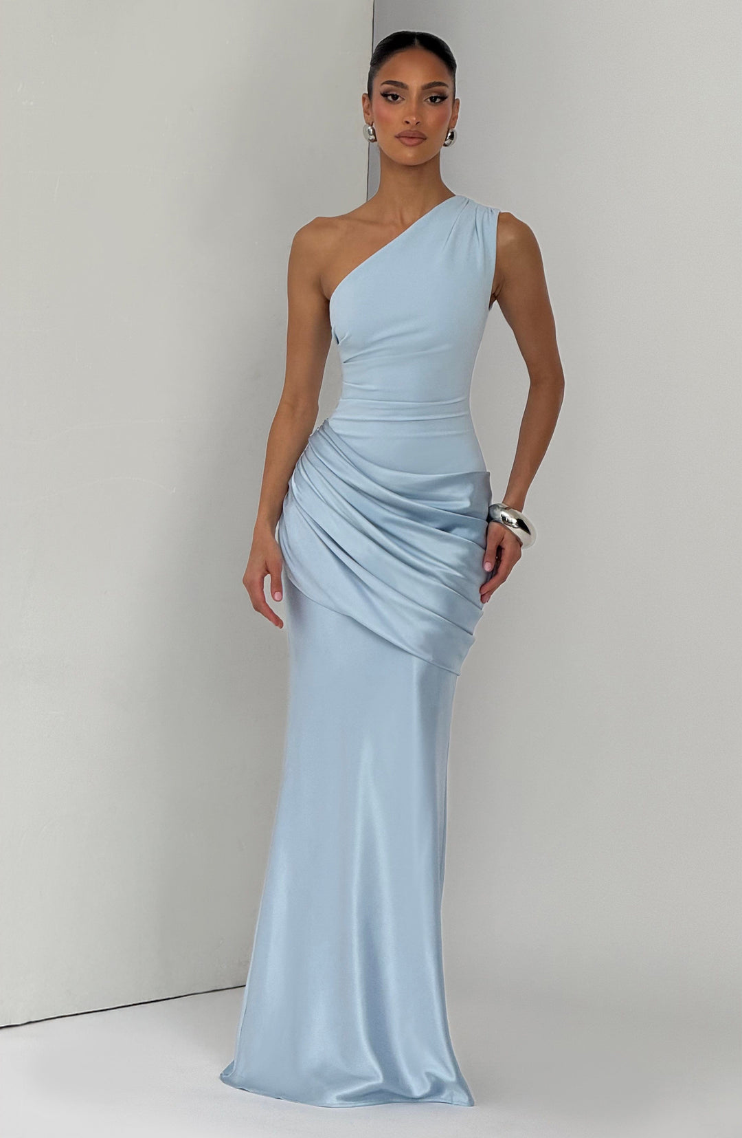 Robe longue bleu clair portée par une mannequin devant un mur gris, buste asymétrique et jupe satinée drapée. 6