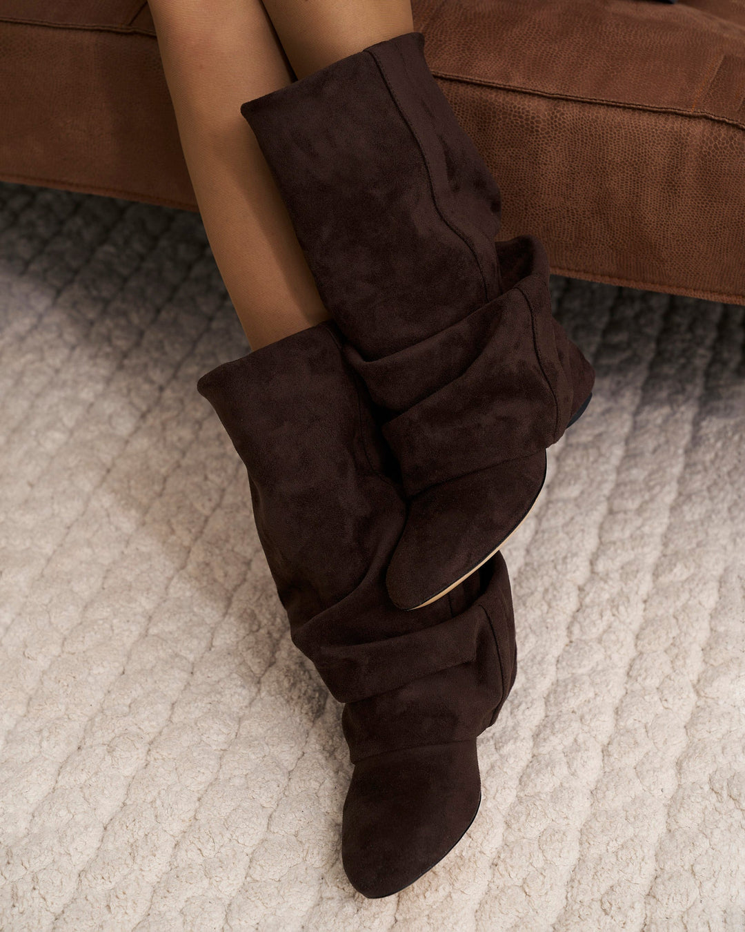 Gros plan sur les bottes chocolat portées aux jambes croisées, assise sur un canapé marron, au-dessus d'un tapis bouclé crème. 2