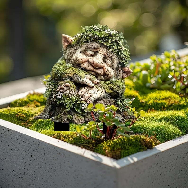 Tuteur d'un troll endormi aux mains croisées et couronne de feuilles, planté dans une jardinière en béton remplie de mousse. 4