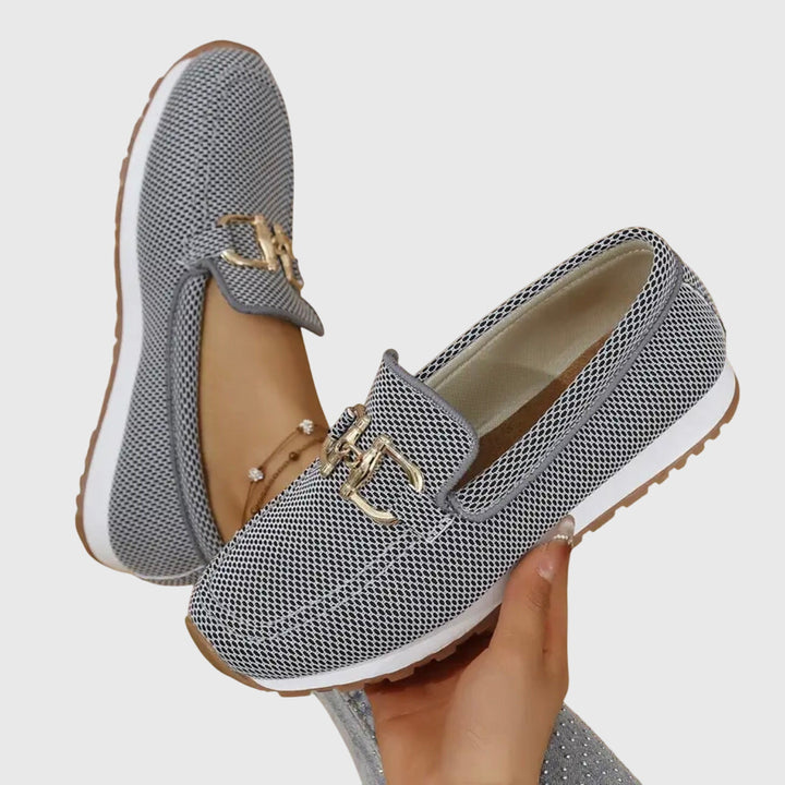 Paire de mocassins gris tenue en main, avec l'intérieur doublé visible et la boucle dorée 8