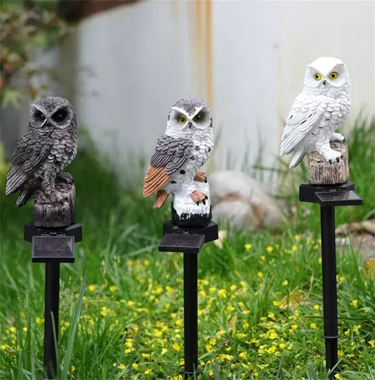Trois hiboux solaires de jour sur leurs piques dans l'herbe, panneaux solaires noirs visibles sous chaque socle en souche. 11