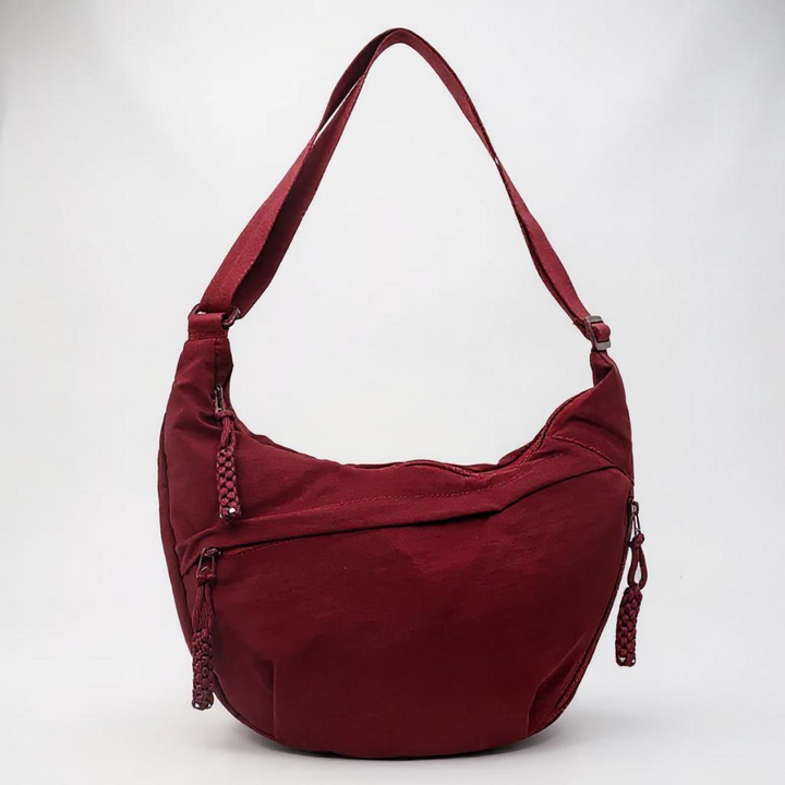 Sac demi-lune bordeaux en nylon vu de face sur fond clair, avec bandoulière réglable et fermetures à glissière à pompons tressés. 1
