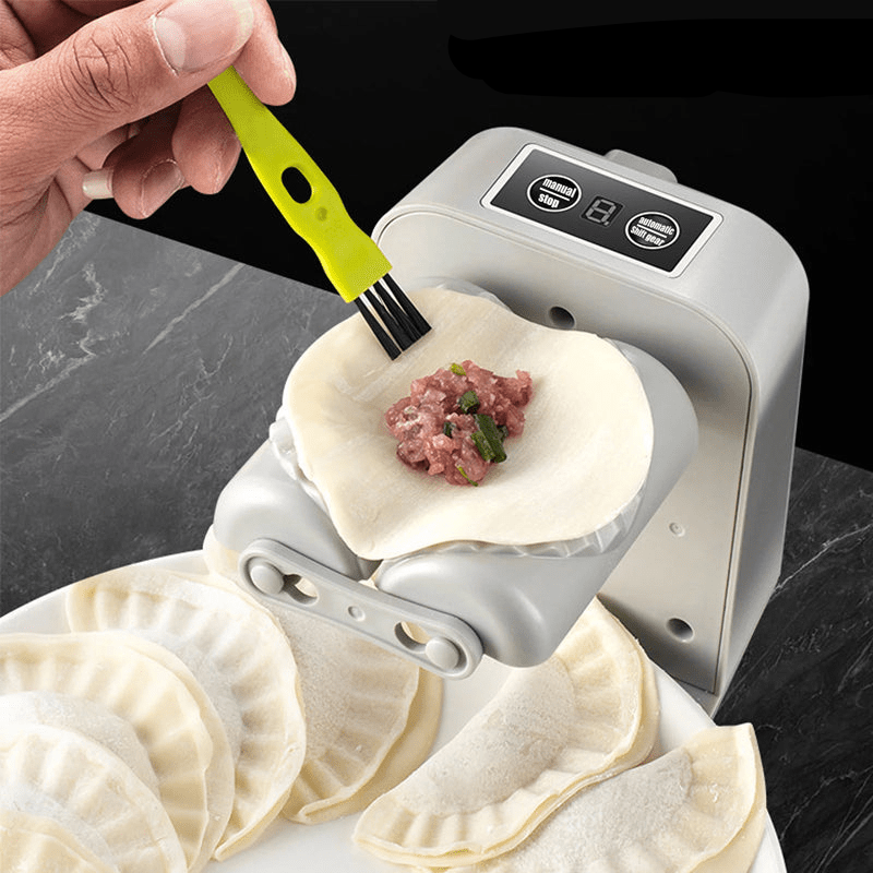 Une main humecte avec un petit pinceau vert le bord d'une pâte garnie de viande hachée sur la machine, raviolis finis au premier plan. 1
