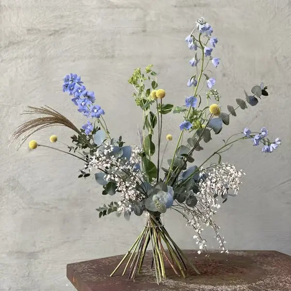 Bouquet aérien de delphiniums bleus, eucalyptus et boutons jaunes, tiges croisées en spirale, sur un support rouillé devant un mur gris. 12