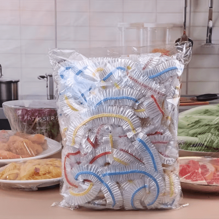 Grand sachet transparent rempli de couvercles élastiques à bandes colorées, posé sur un plan de travail de cuisine devant des plats couverts. 1
