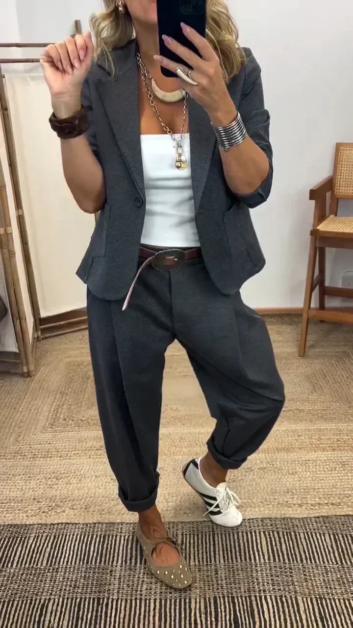 Femme en miroir portant le blazer à un bouton et le pantalon ample gris anthracite, avec haut blanc, ceinture et baskets. 1