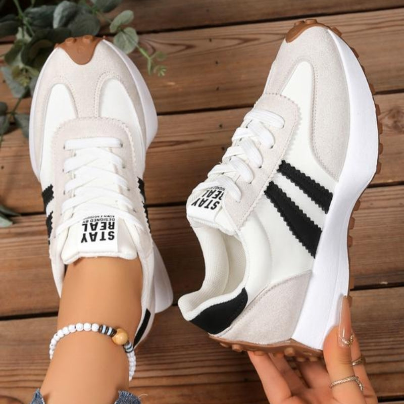 Baskets beige et blanches à bandes noires, une chaussure portée, l'autre tenue en main, sur du bois 10