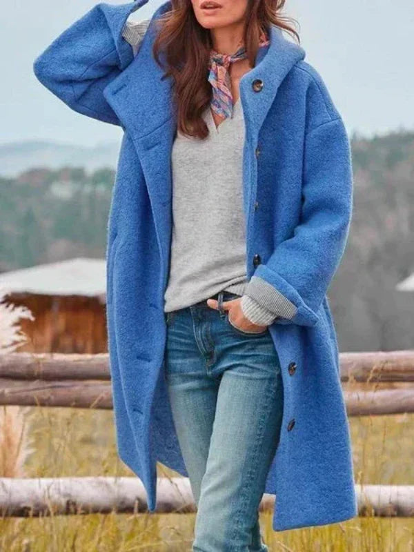 Manteau long à capuche bleu porté ouvert sur un pull gris et un jean, une main dans la poche, devant une clôture. 4