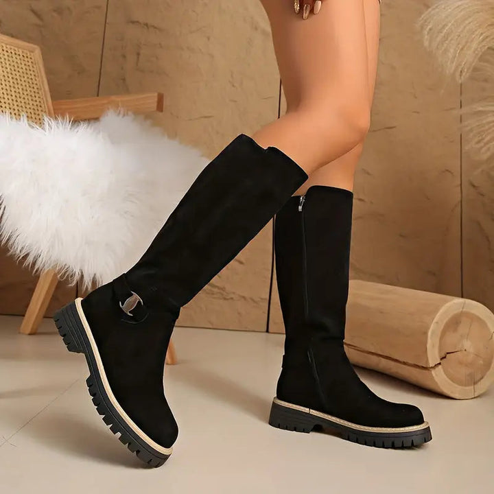 Bottes noires en effet daim portées par une femme, jambe croisée, avec fermeture éclair latérale et boucle métallique dorée. 1