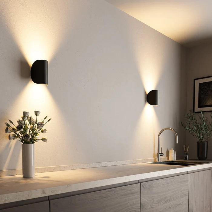 Deux appliques noires diffusant une lumière chaude vers le haut et le bas sur un mur de cuisine avec plan de travail et vase. 1