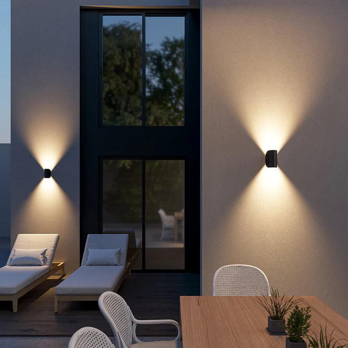 Appliques noires en demi-cylindre éclairant vers le haut et le bas sur le mur d'une terrasse le soir, avec transats et table. 13
