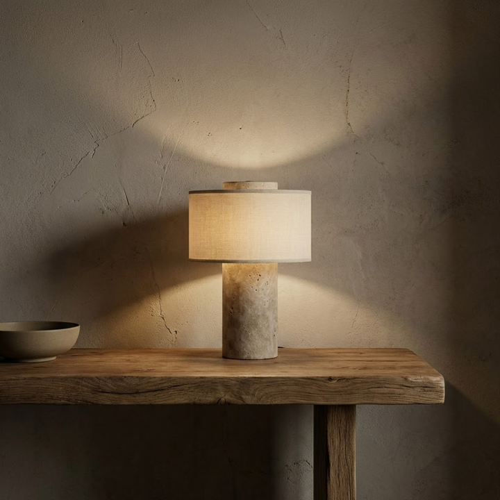 Lampe à pied en pierre poreuse beige allumée sur une console en bois brut devant un mur patiné, avec un bol. 8