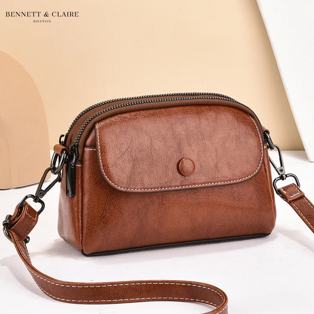 Sac bandoulière marron cognac à rabat avec bouton pression, double zip, surpiqûres blanches et mousquetons noirs, posé sur fond beige. 1