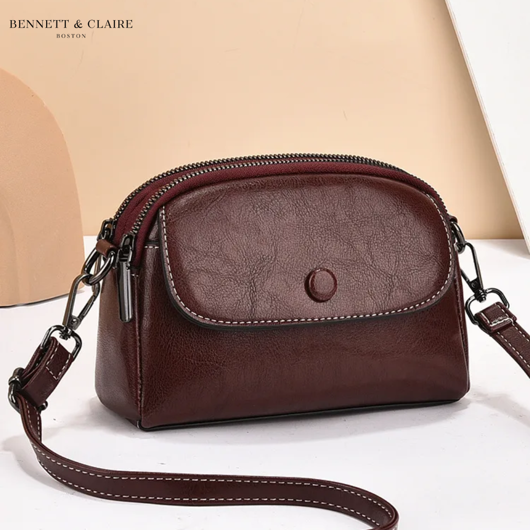 Sac bandoulière bordeaux foncé à rabat avec bouton pression, double zip et surpiqûres blanches, vu de trois quarts sur fond beige. 3
