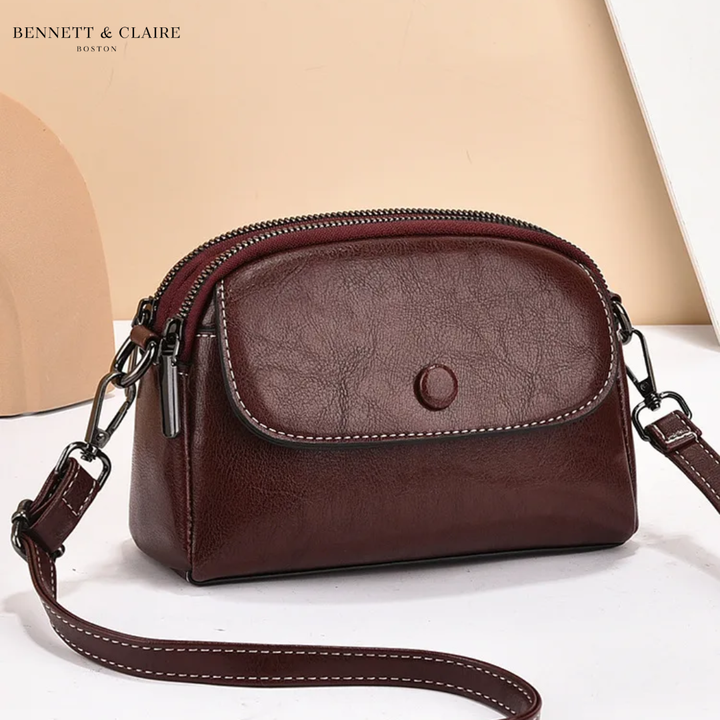 Sac bandoulière bordeaux foncé à rabat avec bouton pression, double zip et surpiqûres blanches, vu de trois quarts sur fond beige. 3