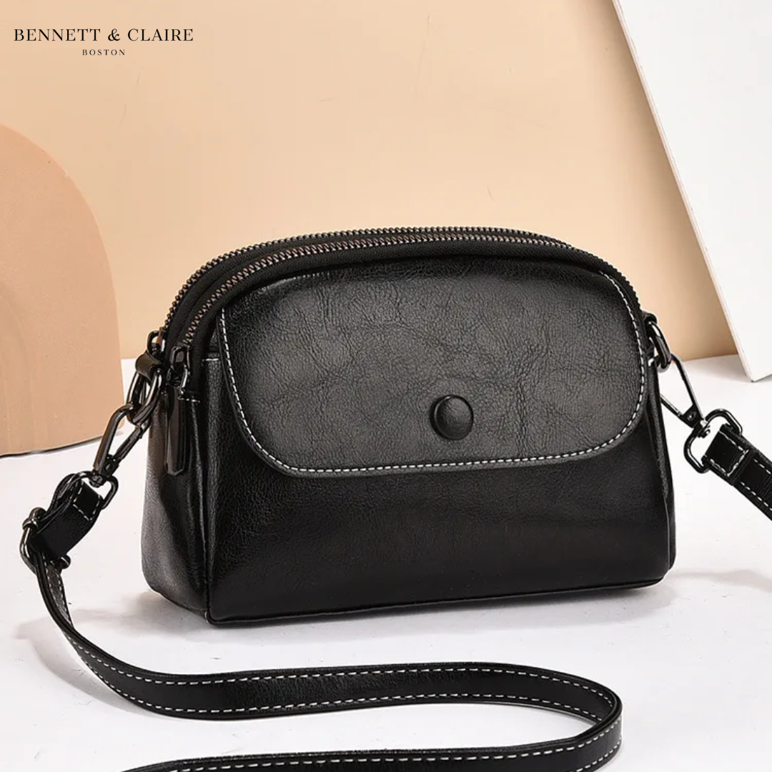 Sac bandoulière noir à rabat avec bouton pression, double fermeture zippée et surpiqûres blanches, bandoulière détachée posée devant. 2