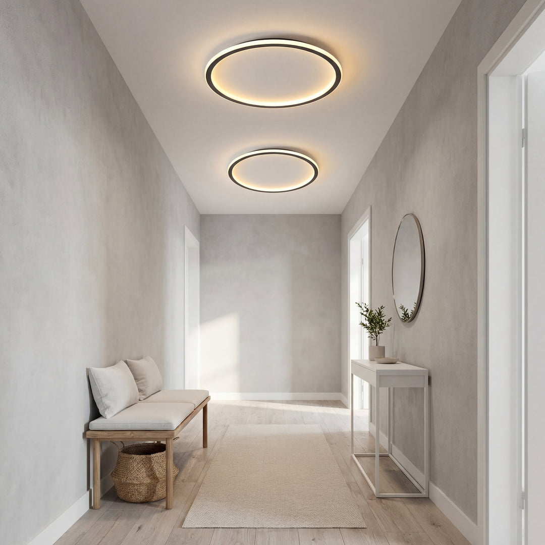 Deux plafonniers en anneau de tailles différentes allumés dans un couloir gris clair, avec banc, console et miroir rond. 6