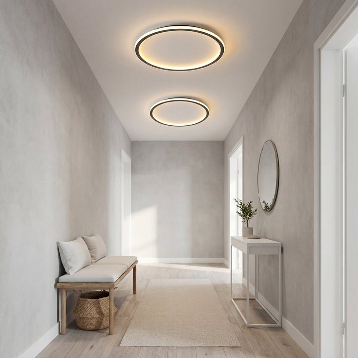 Deux plafonniers en anneau de tailles différentes allumés dans un couloir gris clair, avec banc, console et miroir rond. 6