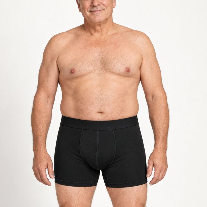 Homme mûr souriant, torse nu, portant un boxer noir à jambe mi-longue, vu de face sur fond blanc. 2