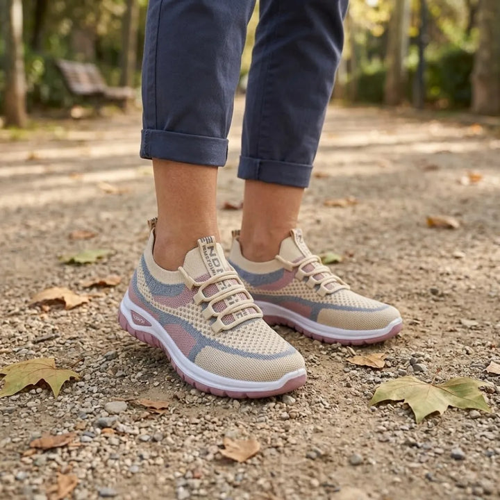 Pieds féminins portant des baskets beige, rose et gris-bleu avec un pantalon bleu retroussé, sur un chemin de parc jonché de feuilles. 29