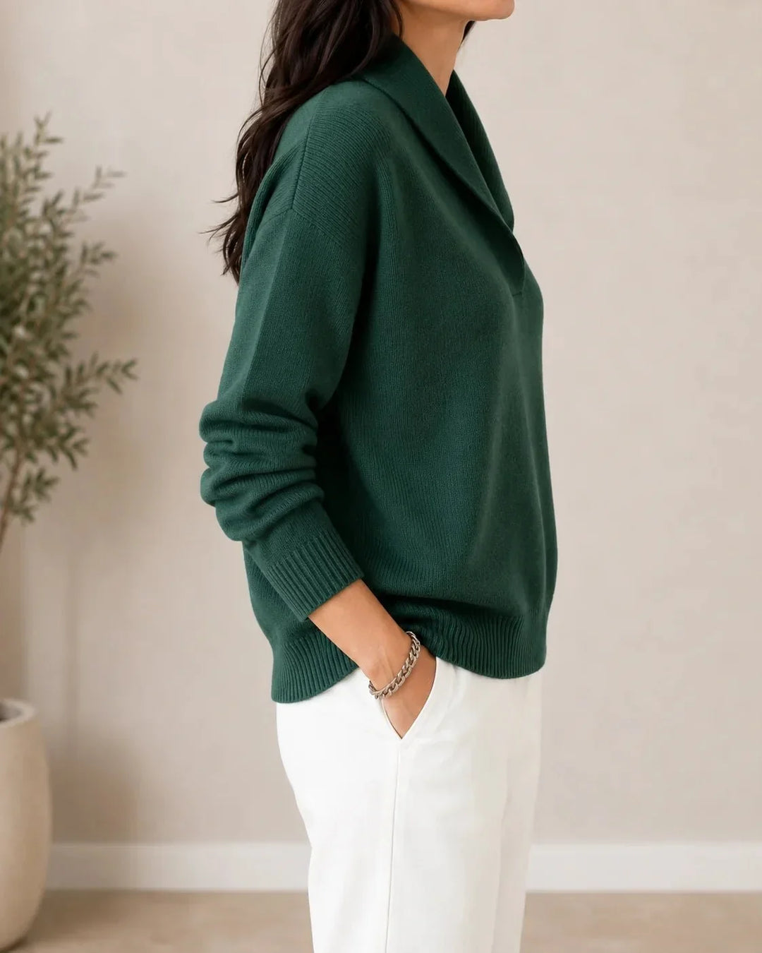 Vue de profil du pull vert foncé porté par un mannequin, main dans la poche, avec plante en arrière-plan. 3